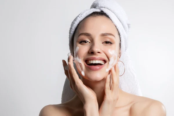 Kobieta w białym ręczniku na głowie myje twarz za pomocą pianki.